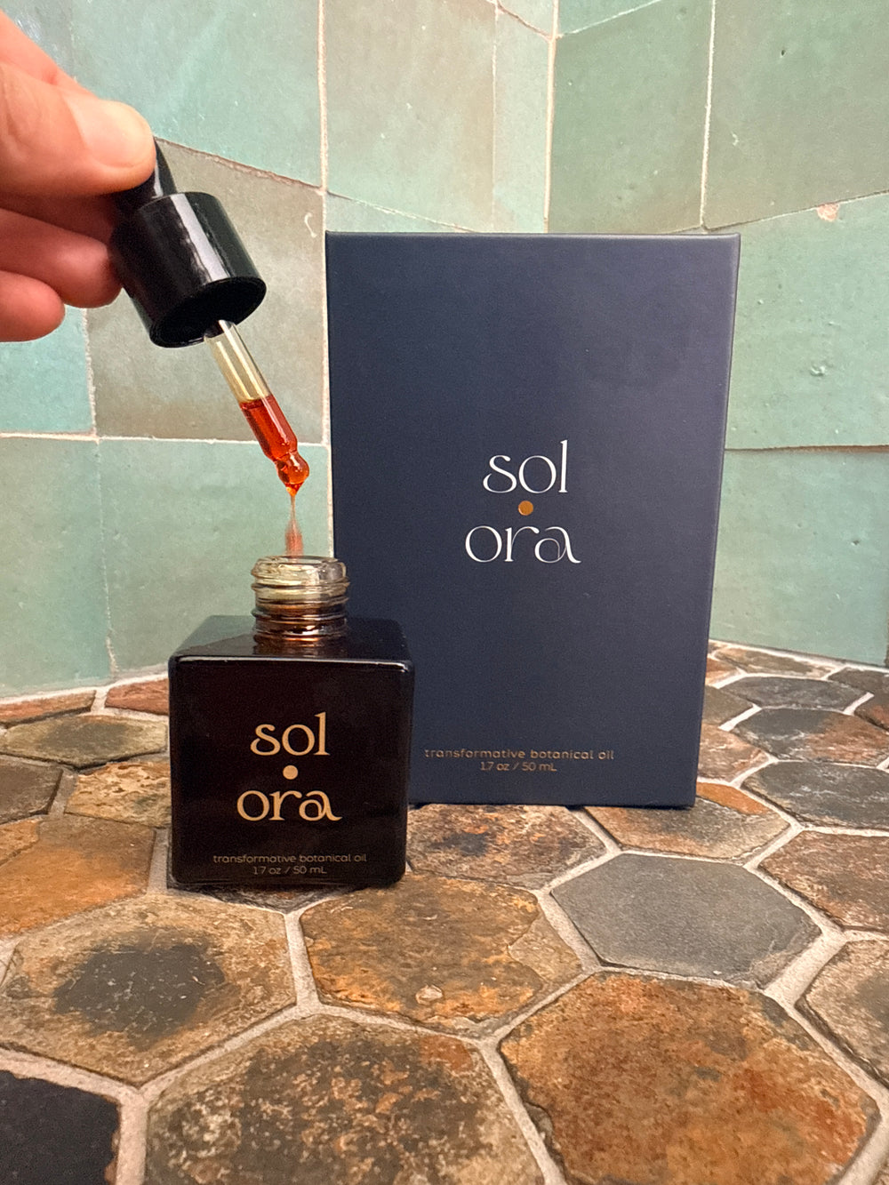 A dropper releasing Sol Ora into the bottle, beside its carton on stone tile