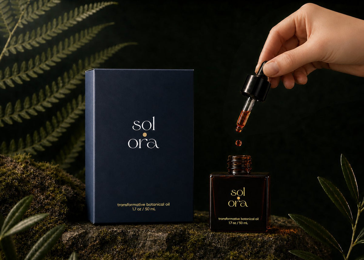 A hand releasing a drop of Sol Ora into the open bottle, beside the carton on moss